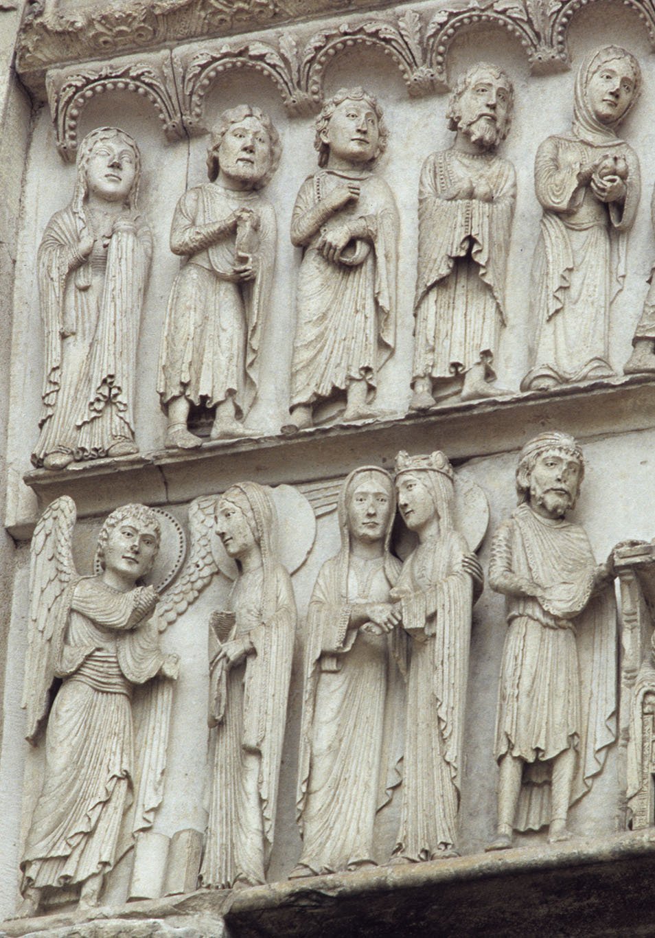 Two rows of delicately sculpted figures in high relief. In the top row, there is a procession of men and women under a decorated arcade. The first two are carrying doves as offerings. In the lower row, we see the Angel Gabriel greeting Mary in the Annunciation scene followed by Mary and Elizabeth in the Visitation scene.