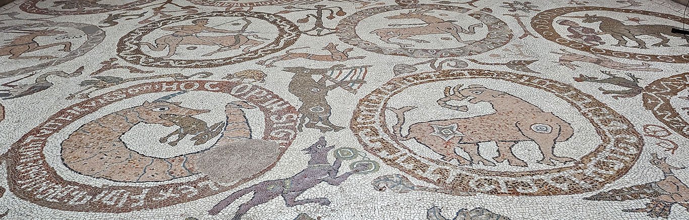 The pavement mosaic is done in earth tones (light and dark browns and grays) against a white background. Large roundels enclose animals and monsters including an elephant, a horned scaly worm-like creature swallowing a goat, and a centaur shooting an arrow at a stag. Between the roundels, a donkey and dog play musical instruments and there are other animals.