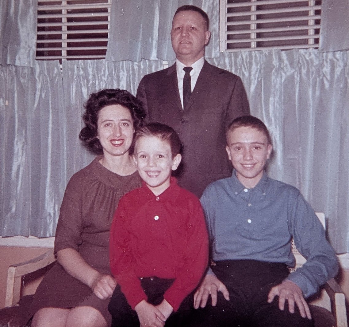 This is a posed family photograph in front of a window with curtains and blinds. My mother and brother sit side-by-side on wooden armchairs. I am between them. In the center, my father stand rigidly behind all of us, wearing a jacket, a white shirt, and thin tie. He has a crewcut.