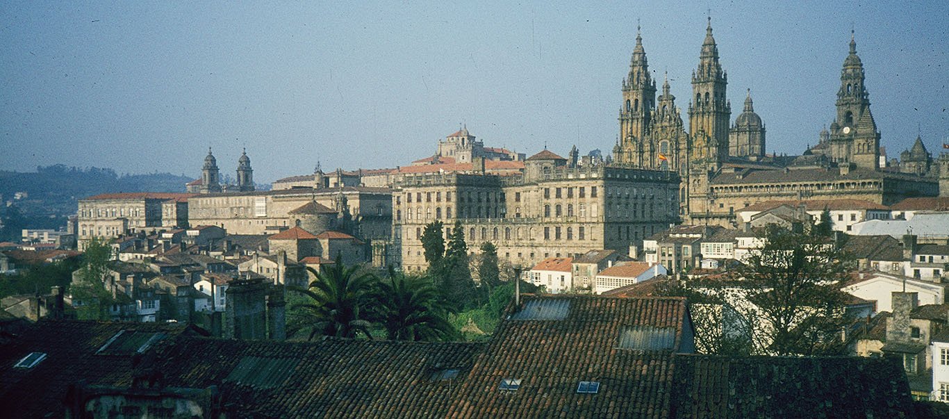 This panoramic view of Santiago de Compostela is dominated by the façade, towers, and cloister buildings of the cathedral. In the foreground, tile roofs lie in the shadows. Beyond that, there is a glimpse of a park with palm trees. Then the major public and ecclesiastical buildings of the center of Santiago rise towards the cathedral with their high walls and towers. Their greyish brown granite is illuminated by the late afternoon sun. They contrast with the smaller group of modest residential houses and buildings with white plastered walls in the lower right.
